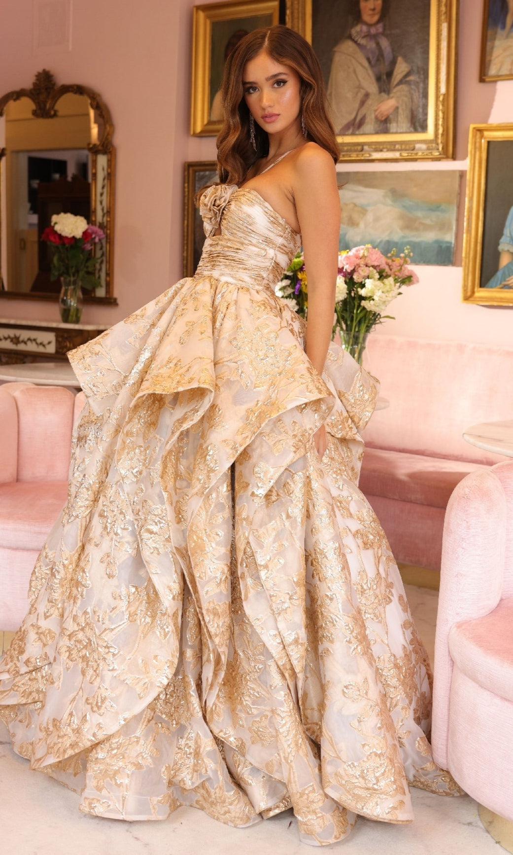 A woman in the Ava Presley 40049 strapless gold and cream embroidered prom ball gown stands in a pink room adorned with 3D flowers and framed portraits.